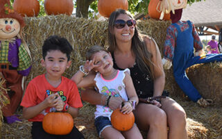 an adult woman and two childen smiling in front of pumpkins